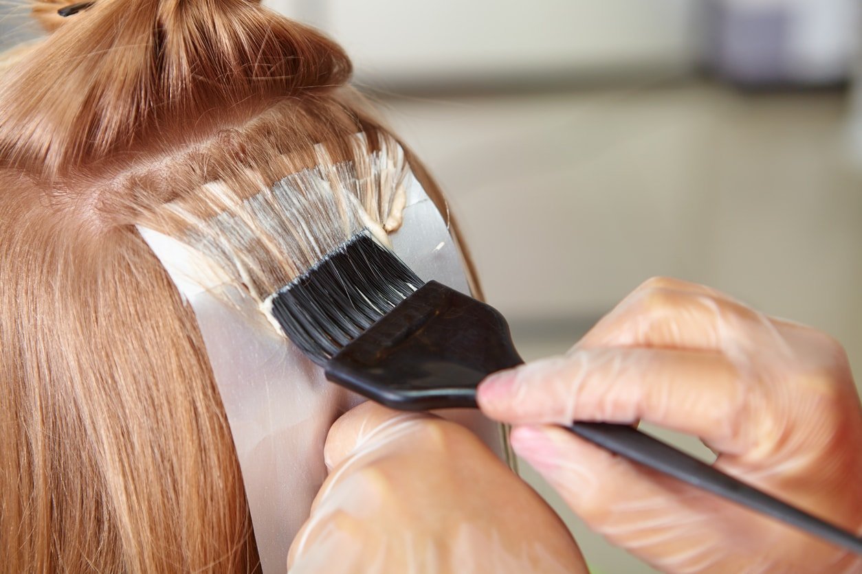 Close-up de mãos pintando mechas no cabelo de uma mulher em um salão de beleza.