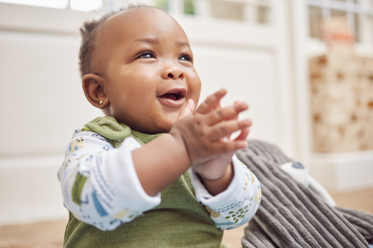 A black baby girl sitting up on the floor clapping her hands together as she smiles happily.