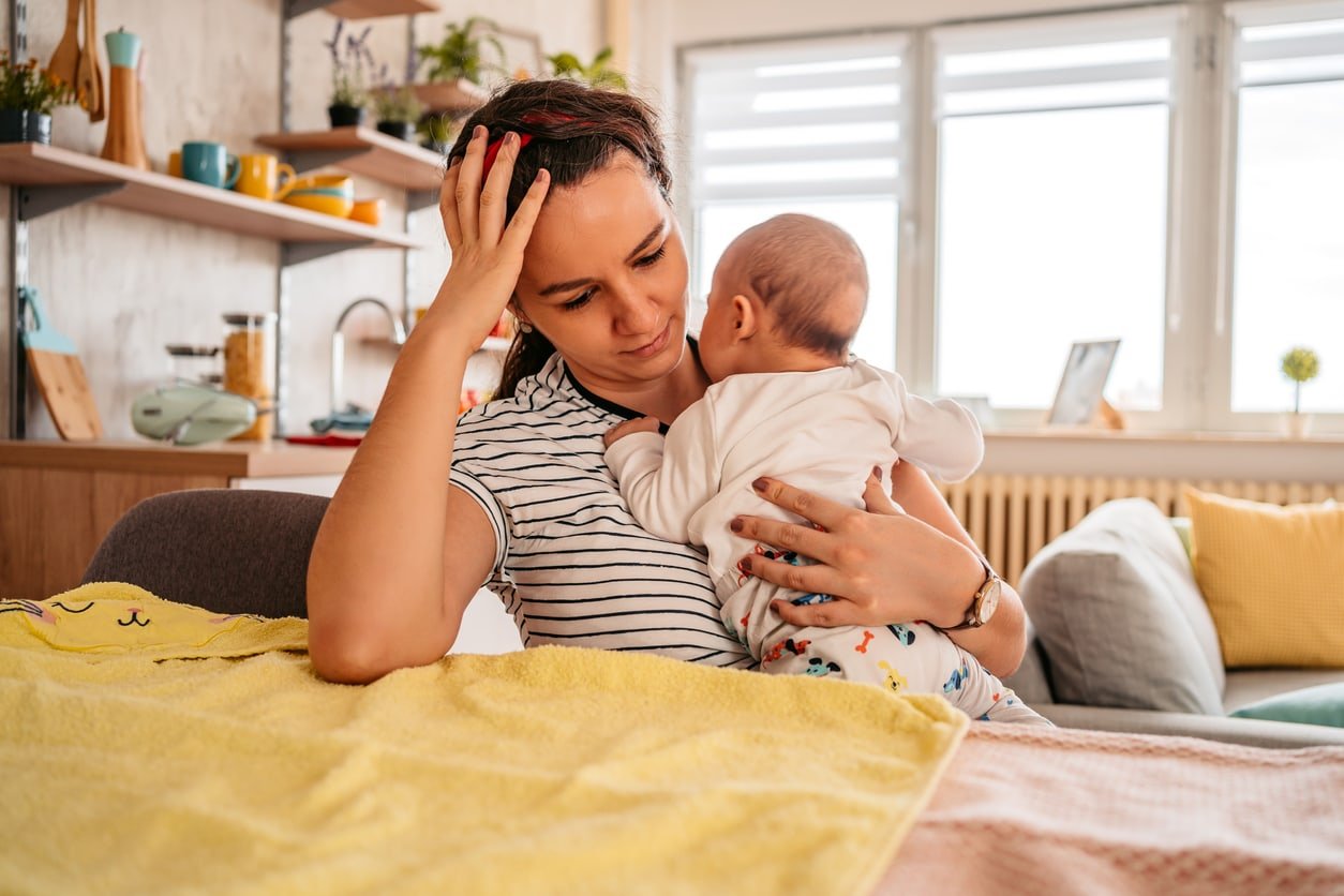 Jovem mãe adulta sentada em uma cadeira em casa, segurando seu filho de três meses, sentindo-se cansada, com sono ou com dor de cabeça.