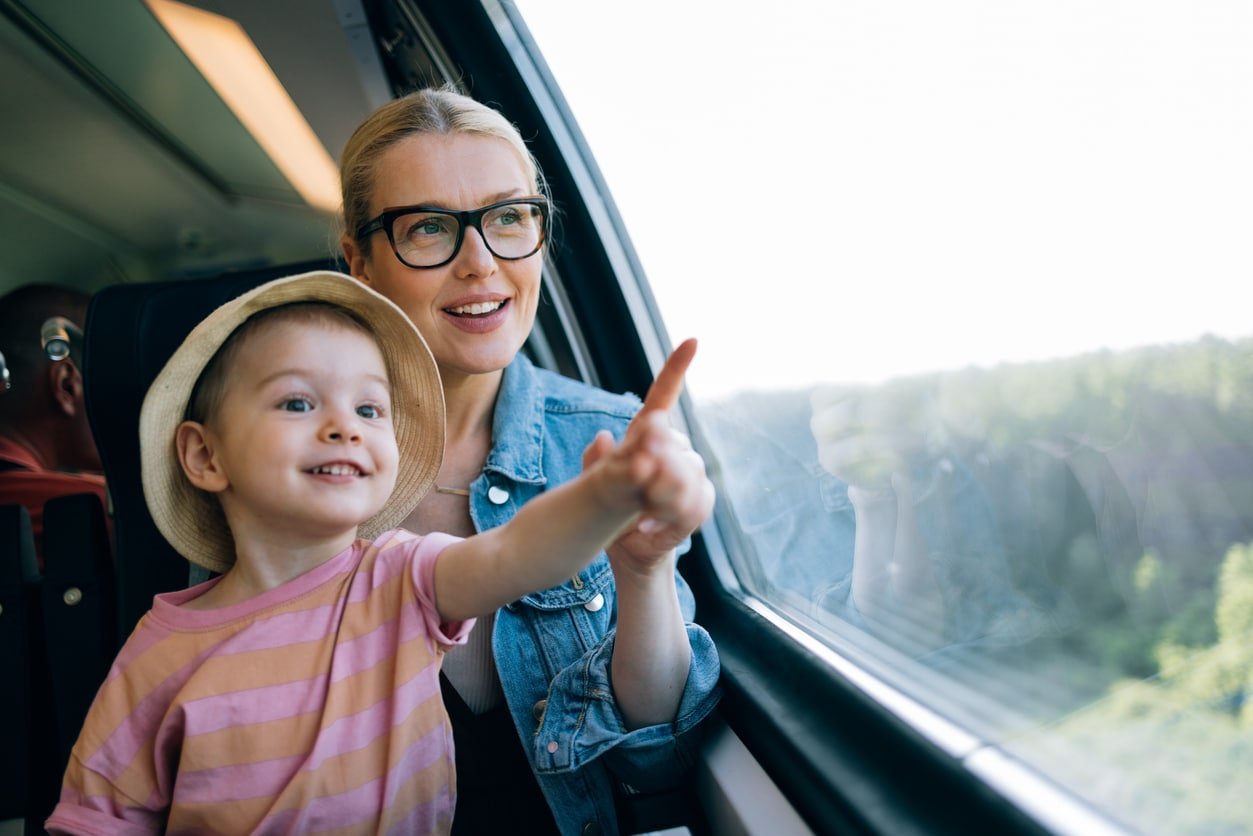 Mãe sorridente mostrando algo para sua filha enquanto está sentada no trem e olhando pela janela.