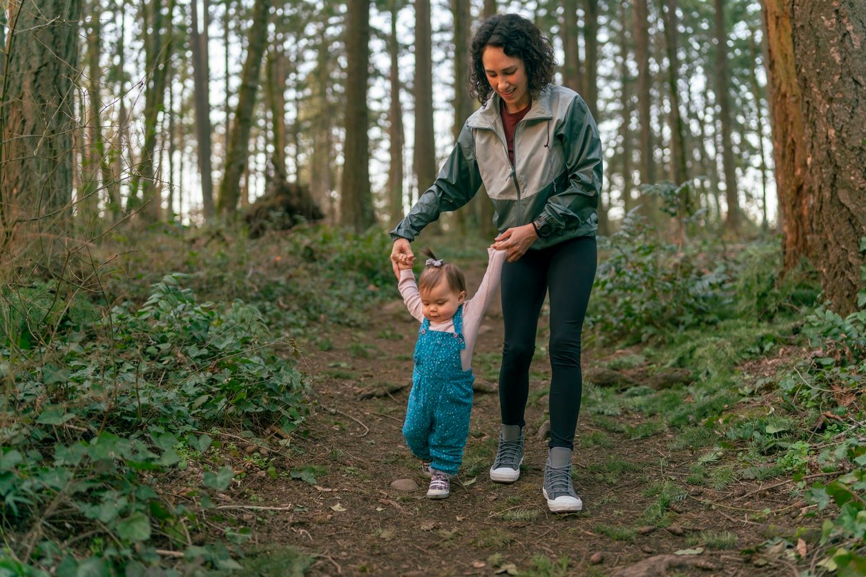 Uma linda mãe mestiça das ilhas do Pacífico está segurando as mãos de sua filha de um ano e ajudando a criança a aprender a andar.  A família está em uma caminhada de aventura na floresta ao pôr do sol.