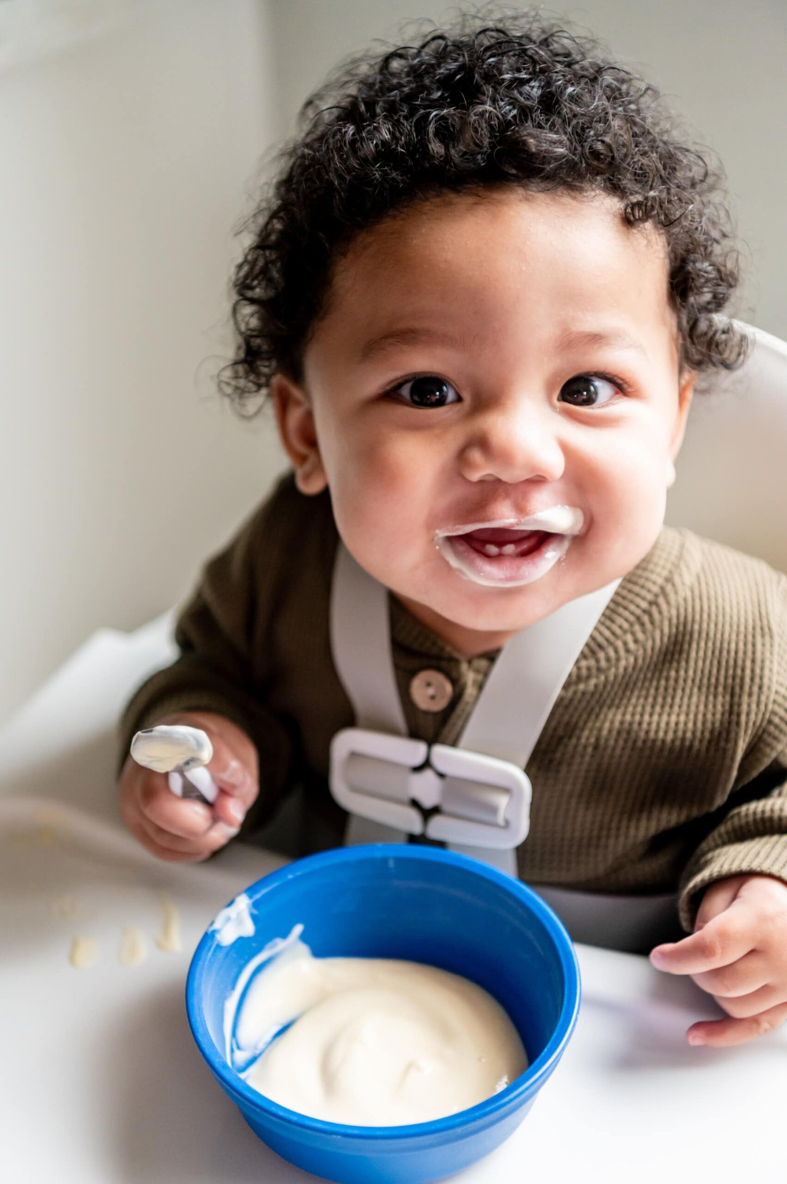 Menino feliz sorrindo em sua cadeira alta, olhando para a câmera.  Ele tem uma tigela de iogurte na bandeja e uma colher com iogurte.