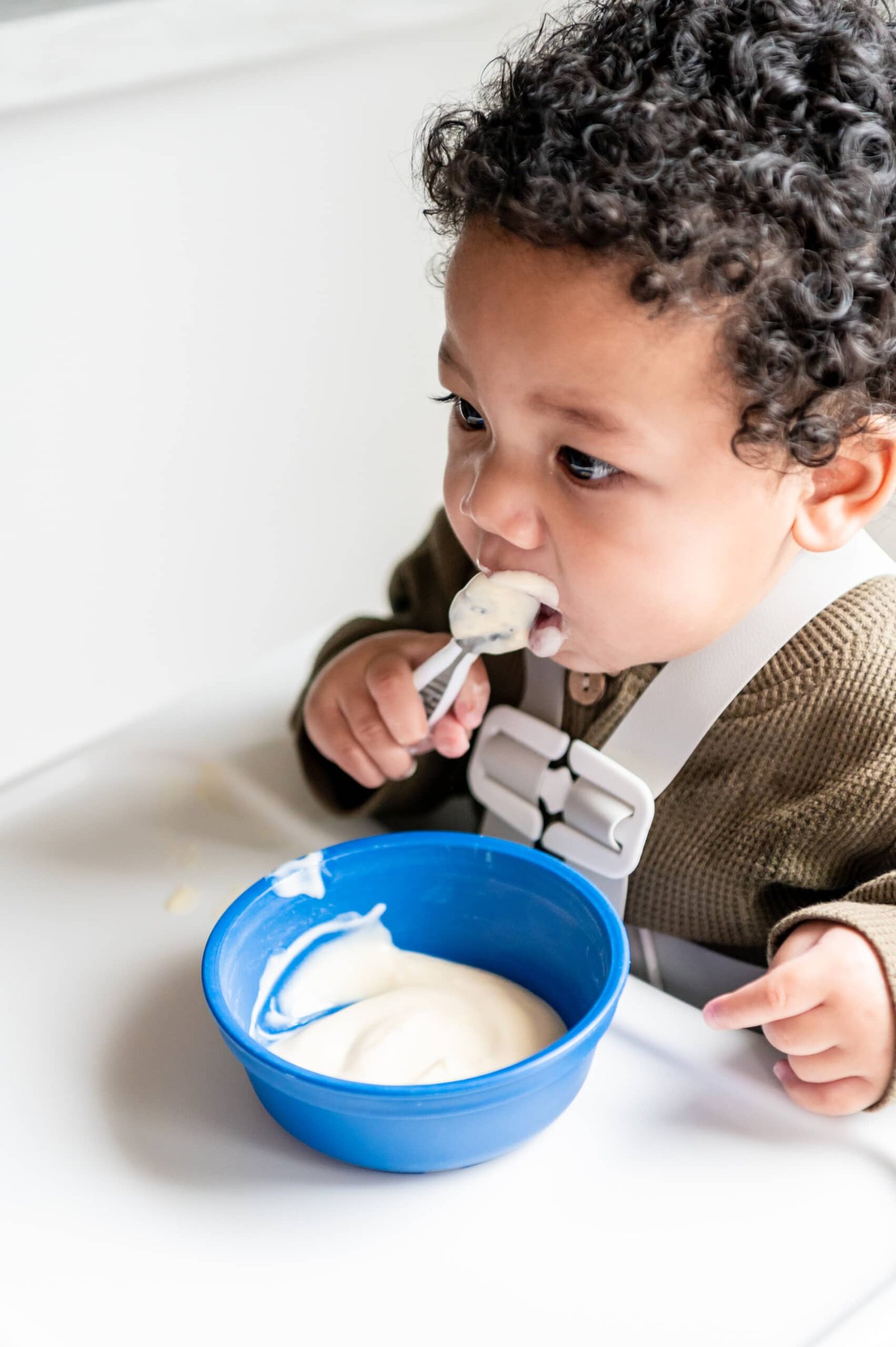 Menino comendo iogurte com uma colher.  Há uma tigela azul com iogurte.