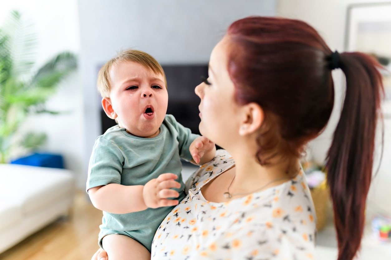 Uma mãe segurando um bebê na sala de estar.  O bebê está doente e com tosse