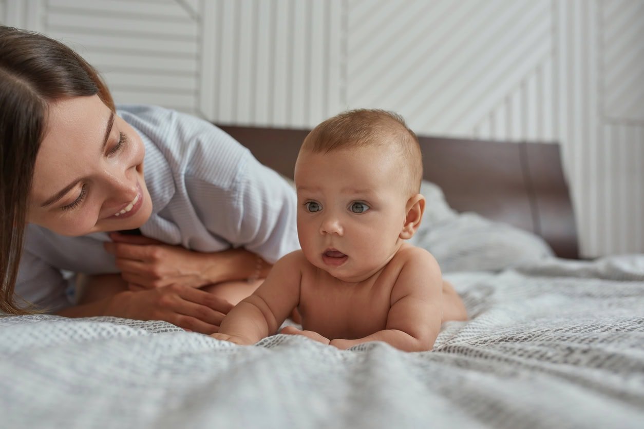 Retrato de criança espantada deitada de bruços perto da mãe sorridente.  Mulher alegre olhando feliz para seu bebê.  Bebê assistindo algo interessante enquanto está de bruços.