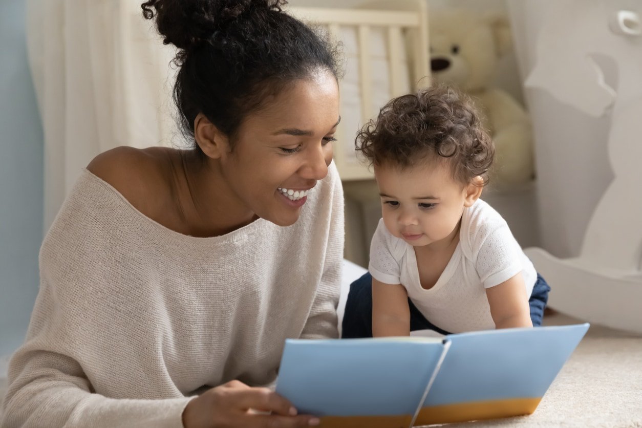Feliz jovem mãe biracial e criança étnica sentam-se no chão no quarto das crianças em casa lendo livro juntos.  Sorridente e amorosa mãe afro-americana se diverte brincando de estudar com um bebê pequeno.
