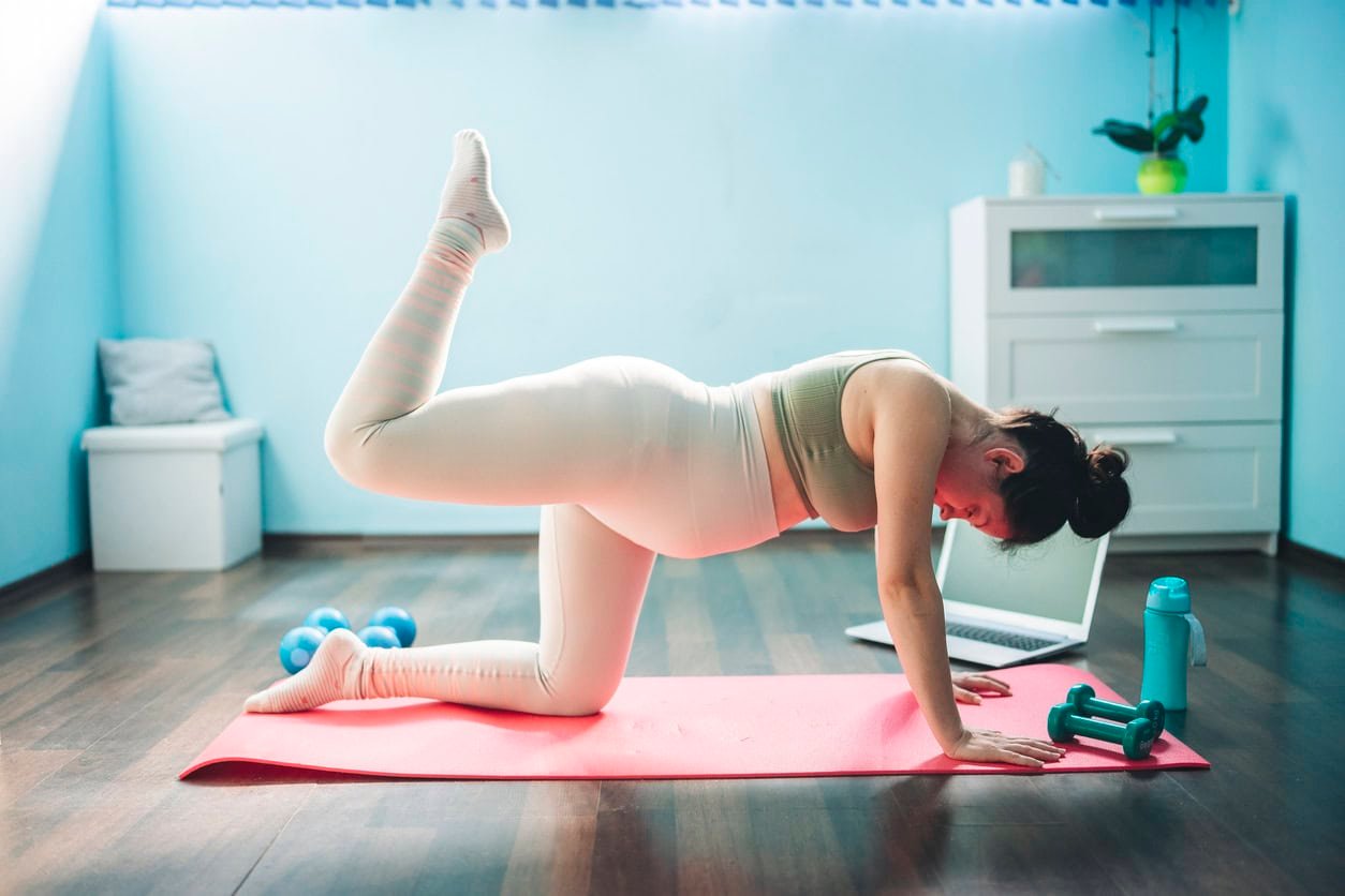 Uma mulher grávida se exercita em um tapete de ioga rosa em uma sala com piso de madeira.  Ela está usando um sutiã esportivo verde e legging branca, levantando uma perna atrás dela.  Perto estão halteres, uma garrafa de água e um laptop.  Um armário e uma planta ficam ao fundo enquanto ela usa hamamélis para aliviar hemorróidas durante a gravidez.