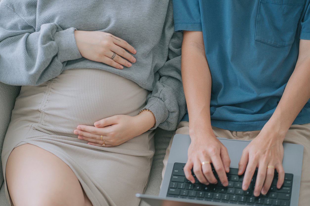 Duas pessoas estão sentadas lado a lado em um sofá.  A pessoa à esquerda está vestindo um suéter cinza e saia de cor clara, com a mão apoiada na barriga de grávida - provavelmente o orçamento do bebê é a prioridade.  A pessoa à direita está com uma camiseta azul, digitando em um laptop.  Ambos usam anéis.