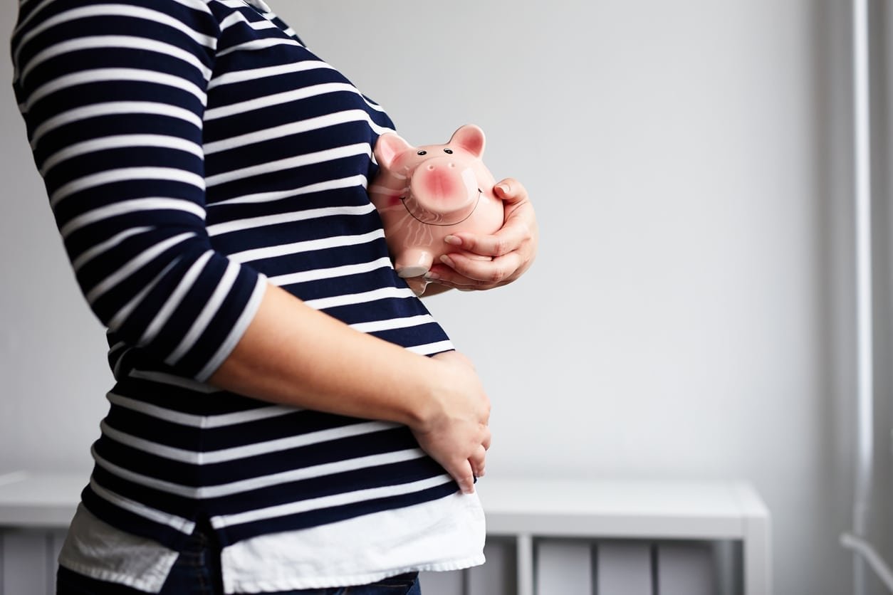 A person wearing a black and white striped shirt, visibly pregnant, holds a pink piggy bank with one hand while resting the other on their stomach. The background is a simple, white-walled room, symbolizing practical saving tips for new moms.