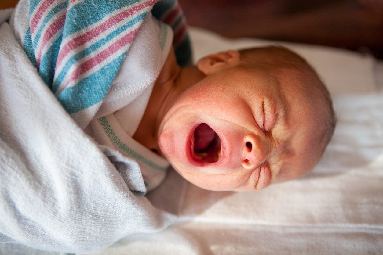 A newborn baby wrapped in a white blanket with teal and pink stripes is lying down and crying, possibly due to a tongue tie. The baby