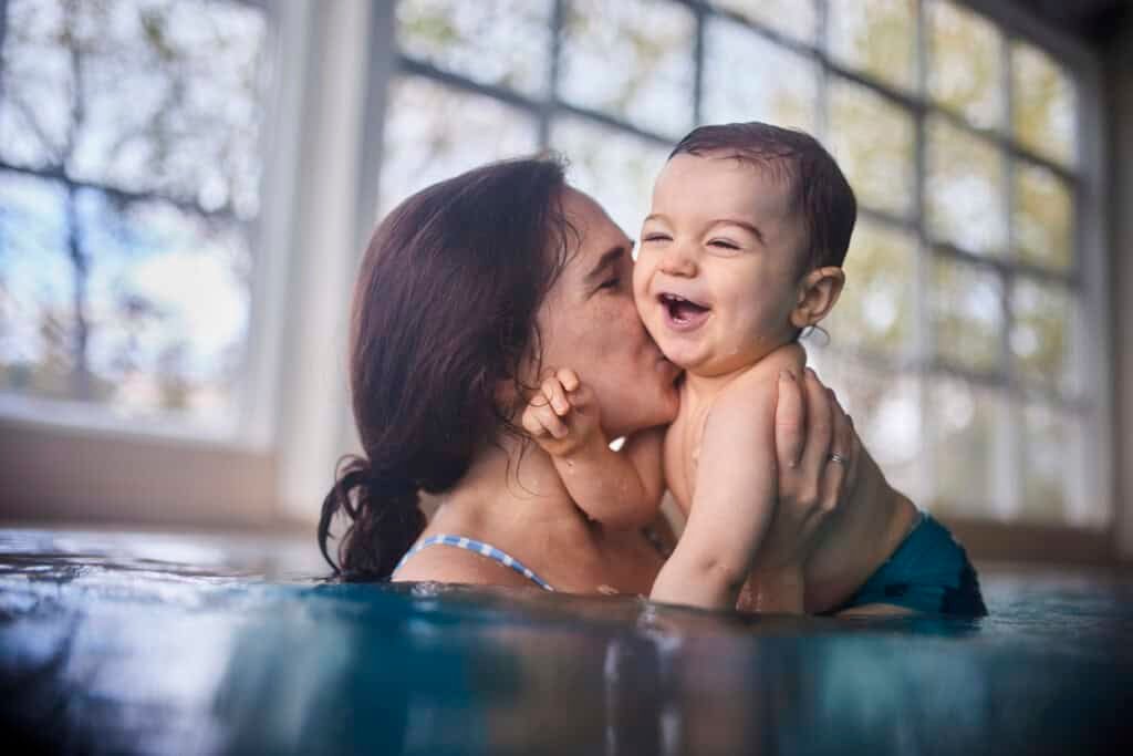 Uma mãe segurando um bebê sorridente em uma piscina aproveita o tempo juntos. Ela beija a bochecha do bebê enquanto a luz natural entra pela parede envidraçada atrás deles. É um momento delicioso, ilustrando a alegria de nadar com bebês e como eles podem aproveitar a água com seus entes queridos.