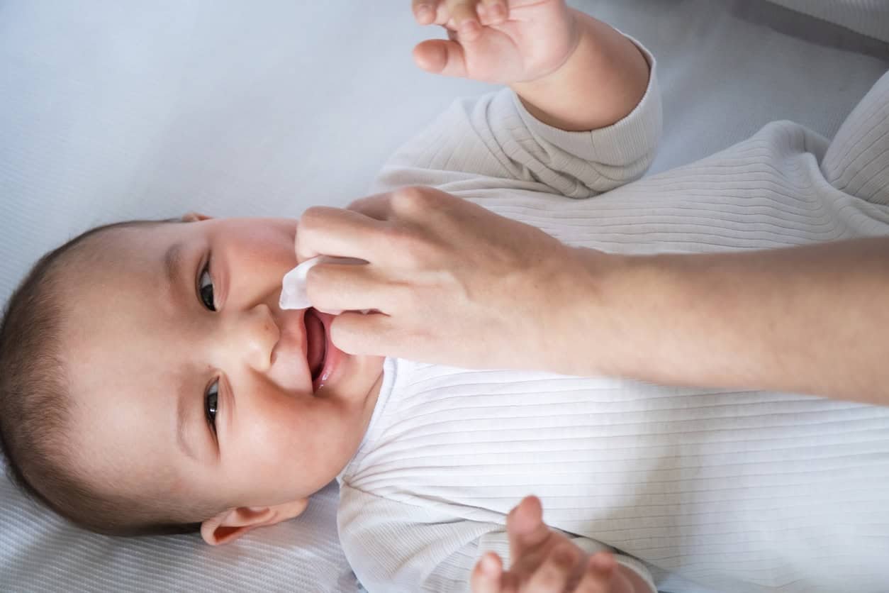 Um bebê está deitado em uma superfície branca vestindo uma roupa branca, enquanto a mão de um adulto limpa gentilmente o nariz do bebê com um lenço. O bebê, olhando para cima e sorrindo, apesar de cuspir mais do que o normal, parece contente e à vontade.