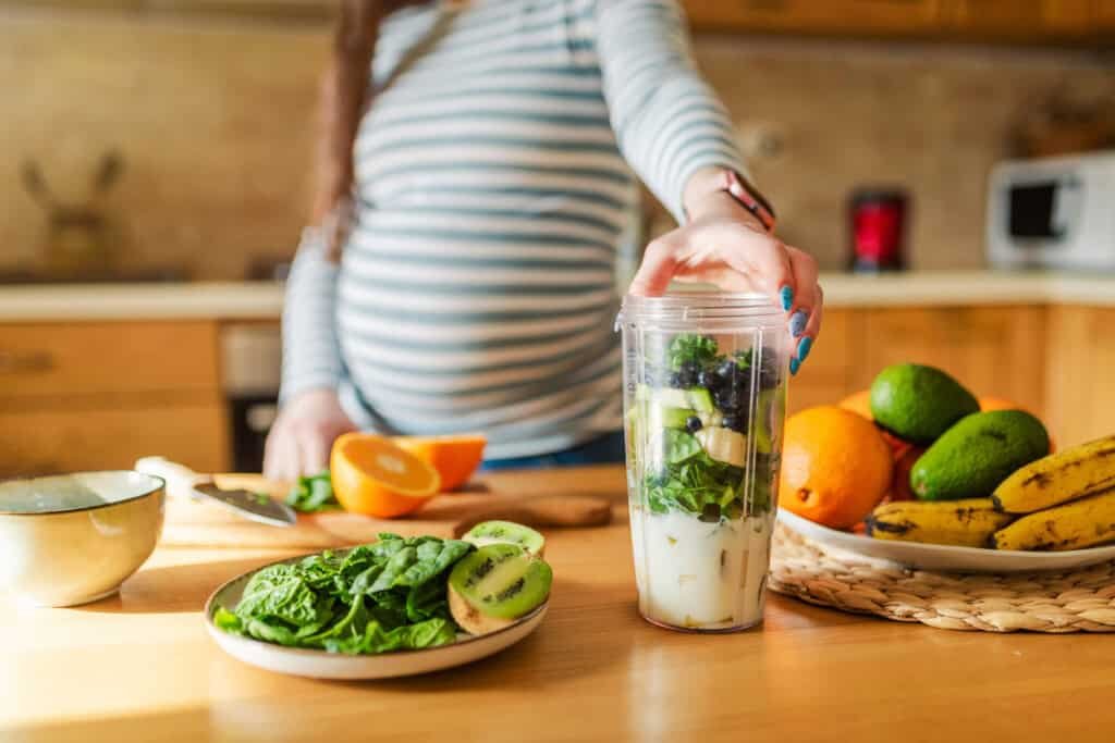 Uma mulher grávida com uma blusa listrada, consciente do inchaço na gravidez, prepara um smoothie em um copo de liquidificador no balcão da cozinha. Várias frutas, incluindo kiwi, abacate, banana e espinafre estão dispostas nas proximidades, junto com uma faca e uma tigela.