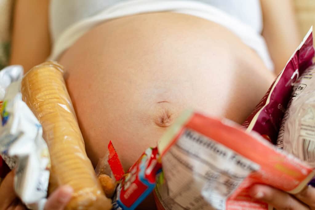 Close de uma barriga de grávida com vários lanches descansando sobre ela, incluindo biscoitos, salgadinhos e outros alimentos embalados. A imagem destaca a parte central de uma pessoa deitada, sutilmente sugerindo a experiência comum de pés inchados durante a gravidez. Uma mulher grávida segurando muitos lanches e desejos por comida em volta da barriga do bebê.