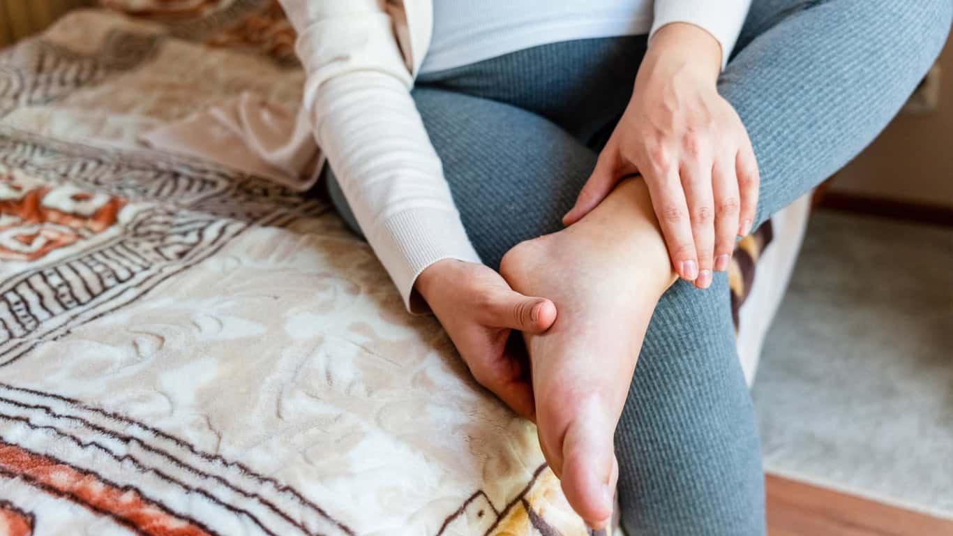 A pregnant woman sitting on the edge of a bed is holding her left foot with both hands, perhaps due to swelling in pregnancy. They are wearing grey leggings and a light-colored top. The bed has a patterned blanket.
