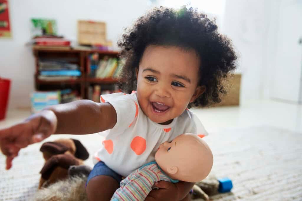 Uma jovem com cabelos cacheados está sentada no chão, sorrindo e apontando para longe. Elas estão segurando uma boneca. Ao fundo, há uma estante com livros e brinquedos espalhados, perfeita para várias atividades para crianças de 18 meses. O quarto é bem iluminado e parece ser uma sala de jogos.