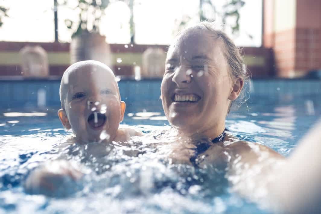 An adult and a child are smiling and splashing in a swimming pool. The adult is holding the child close as they enjoy the water together, making it a delightful baby swimming experience. The background shows a blurred indoor pool area with sunlight streaming in.