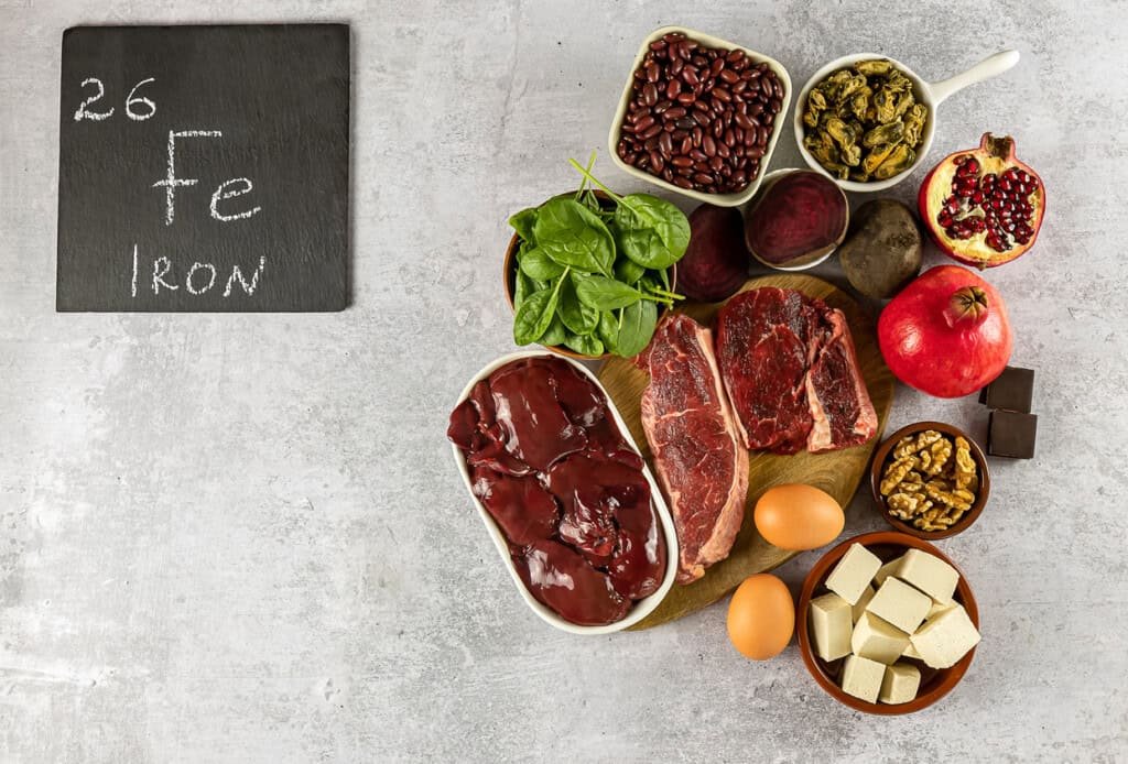 Uma variedade de alimentos ricos em ferro são dispostos em uma superfície cinza claro, incluindo bifes de carne, fígado, espinafre, feijão, nozes, ovos, tofu, romã, beterraba e chocolate amargo. Perfeito para tratar anemia na gravidez. Um quadro negro exibindo 