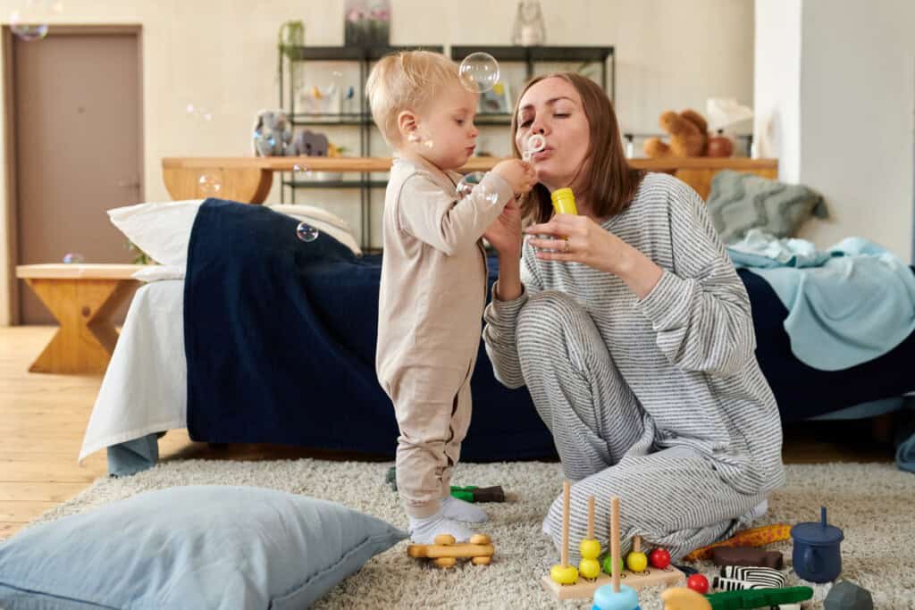 Uma criança pequena e um adulto de pijama sopram bolhas em um quarto. O de 17 meses está de pé enquanto o adulto está ajoelhado no chão. O quarto tem brinquedos espalhados, uma cama com um cobertor azul escuro e uma prateleira de madeira ao fundo. O momento lúdico deles se encaixa perfeitamente no cronograma de sono do bebê de 17 meses.