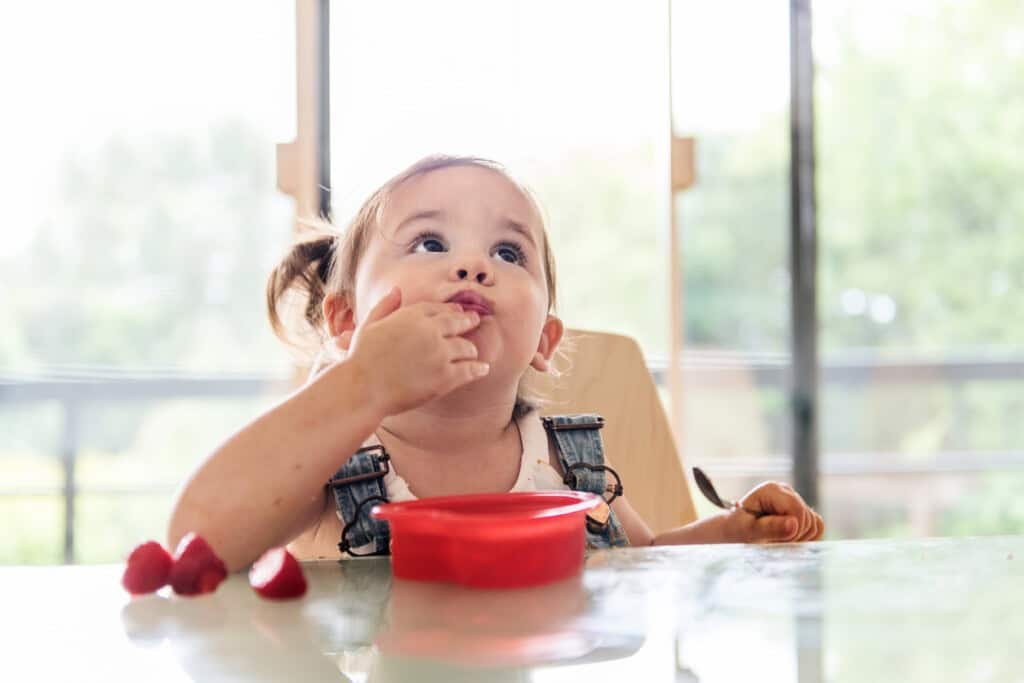 Uma menina com rabo de cavalo está sentada à mesa, comendo de uma tigela vermelha. A criança, provavelmente passando por uma regressão do sono de 18 meses, está vestindo uma camisa branca com macacão jeans e segurando uma colher. Vários morangos estão sobre a mesa. O fundo mostra uma janela brilhante com vegetação do lado de fora.