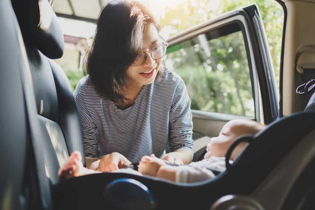 Uma mulher asiática com óculos e uma camisa listrada coloca um bebê em uma cadeirinha de carro, preparando-se para uma viagem de carro com o bebê. A porta do carro está aberta, e a luz do sol filtra através das árvores ao fundo.
