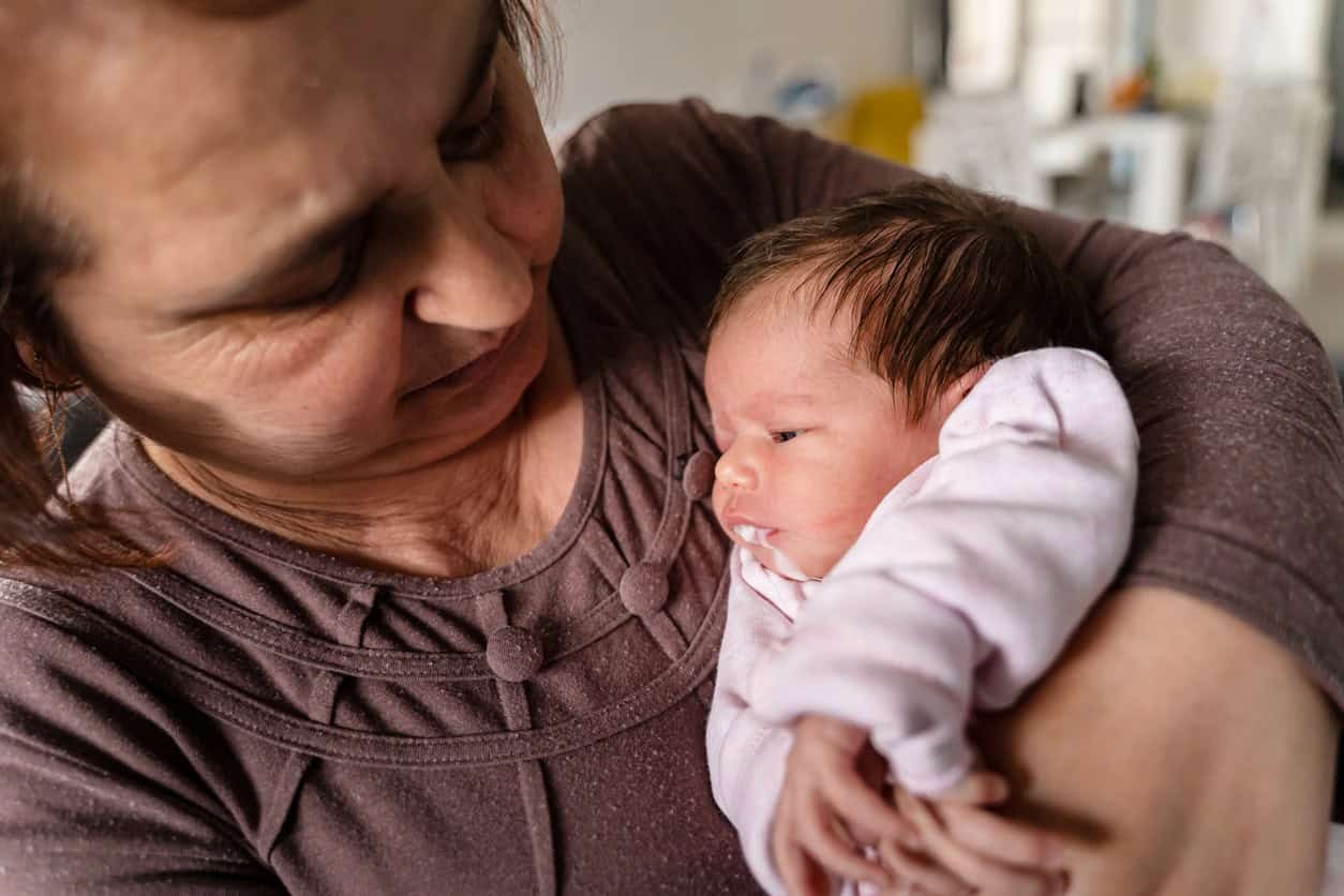Uma mulher com um suéter marrom segura um bebê recém-nascido enrolado em um cobertor rosa.  Ela parece ser uma avó.  Ela está olhando para o bebê com uma expressão gentil, aparentemente despreocupada com possíveis ocorrências de o bebê cuspir mais do que o normal.  O fundo parece ser um ambiente interno com alguns móveis fora de foco.