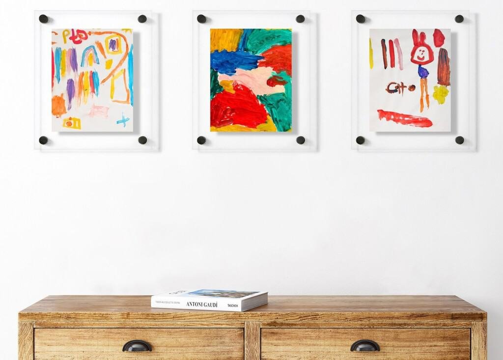 Três pinturas abstratas coloridas em molduras brancas são montadas em uma parede branca acima de um armário de madeira com duas gavetas. Um livro com o título ANDREAGIO é colocado no armário. As pinturas apresentam cores vibrantes e desenhos infantis, reminiscentes de obras de arte infantis caprichosas.