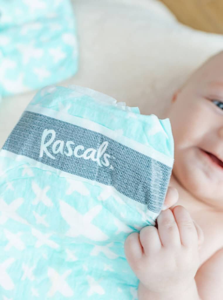 Um bebê segura uma fralda turquesa estampada com desenhos brancos e a palavra Rascals escrita na cintura. Os olhos do bebê e parte do seu rosto são visíveis, com foco nas Fraldas Rascals Premium em primeiro plano.