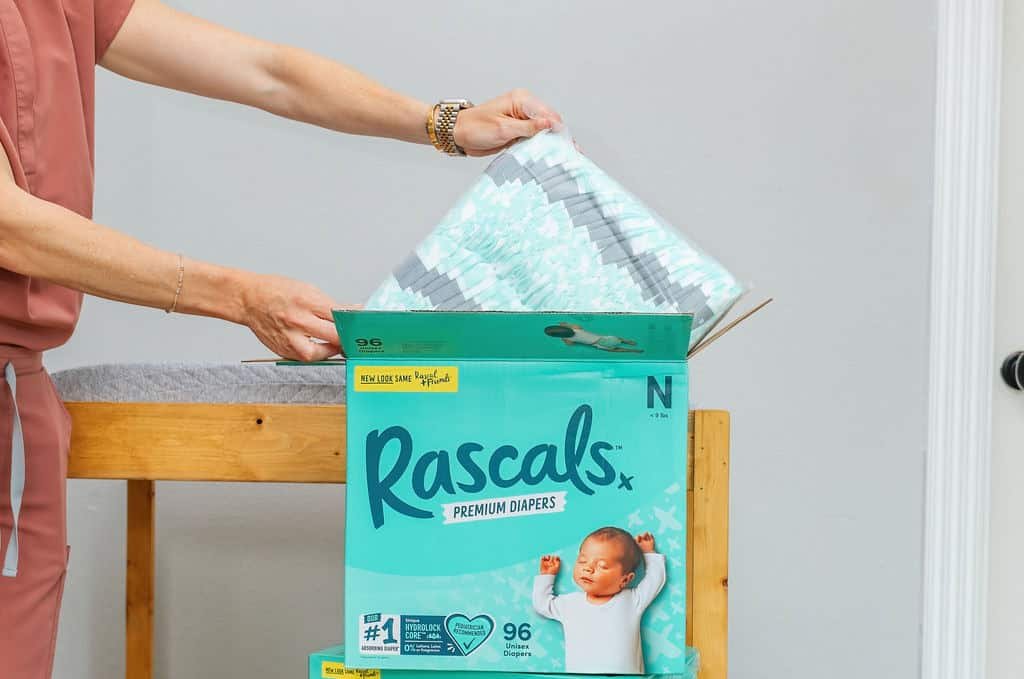 Uma pessoa está abrindo uma caixa de Rascals Premium Diapers. Ela está tirando um pacote de fraldas da caixa verde, que apresenta um bebê dormindo e as palavras Rascals Premium Diapers, tudo isso enquanto está em uma mesa de troca de madeira.