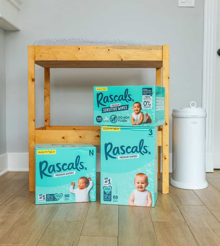 Uma mesa de troca de madeira com uma almofada cinza em cima é colocada contra uma parede cinza claro. Embaixo, há três caixas de fraldas Rascals Premium nos tamanhos N, 1 e 3, junto com uma caixa de lenços umedecidos Rascals sensitive. Um balde de fraldas branco está ao lado da mesa.