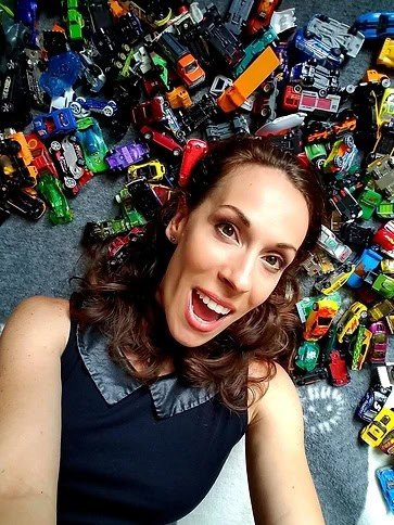 Tara Clark está deitada no chão cercada por vários carrinhos de brinquedo. Ela está sorrindo amplamente e parece estar tirando uma selfie. Ela tem cabelo cacheado e está usando uma blusa preta sem mangas com gola. Os carrinhos de brinquedo são de várias cores e tipos.