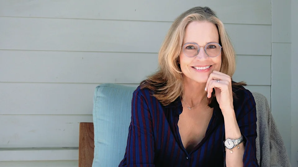Janet Lansbury, usando óculos e vestida com uma camisa azul escura com listras vermelhas, está sentada em uma cadeira de cor clara. Ela tem a mão esquerda apoiada no queixo e está sorrindo para a câmera. Ela está usando um relógio no pulso esquerdo.