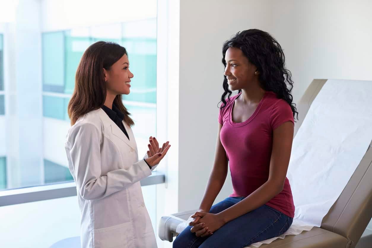 A doctor in a white lab coat is speaking with a woman seated on an examination table. The woman, wearing a pink shirt and jeans, is smiling and listening attentively. The setting appears to be a medical office with a window in the background, as if shes just received good news from her letter to OB.