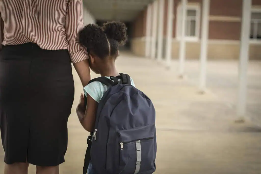 Uma criança com uma grande mochila azul dá as mãos a uma mulher adulta, vista por trás. Eles estão andando sob uma passarela coberta ao lado de um prédio. O adulto veste saia preta e camisa listrada, enquanto a criança veste uma camisa de cor clara - coisas sobre as quais nunca provocar seu filho podem moldar momentos tão preciosos para sempre.