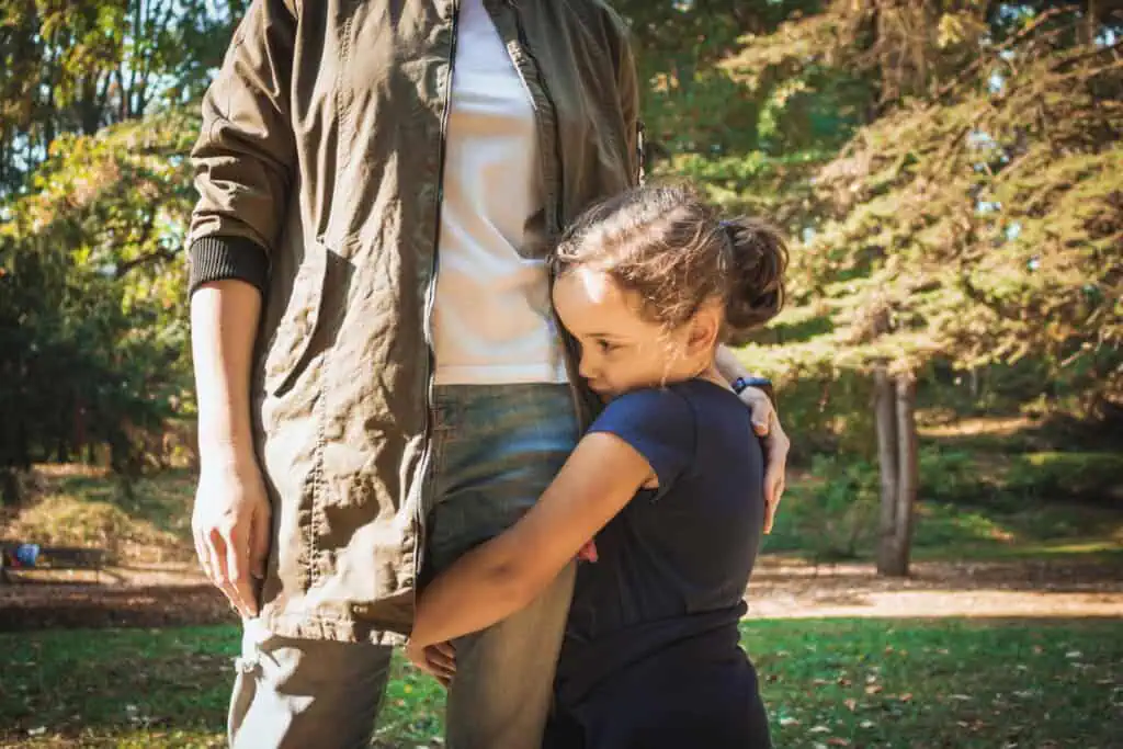 Uma criança de cabelos cacheados abraça a perna de um adulto ao ar livre, um lembrete do vínculo que deve ser nutrido com cuidado. O adulto veste jaqueta verde e calça jeans, e a criança, de camisa azul marinho, encontra conforto neste espaço arborizado, com a luz do sol filtrada pelas árvores.