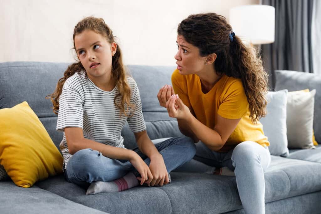 Uma mulher de camisa amarela está sentada em um sofá, conversando animadamente com uma jovem de camisa listrada. A menina, sentada de pernas cruzadas e revirando os olhos, parece desinteressada ou irritada. Parece que eles estão enfrentando a luta clássica de como lidar com a conversa fiada.