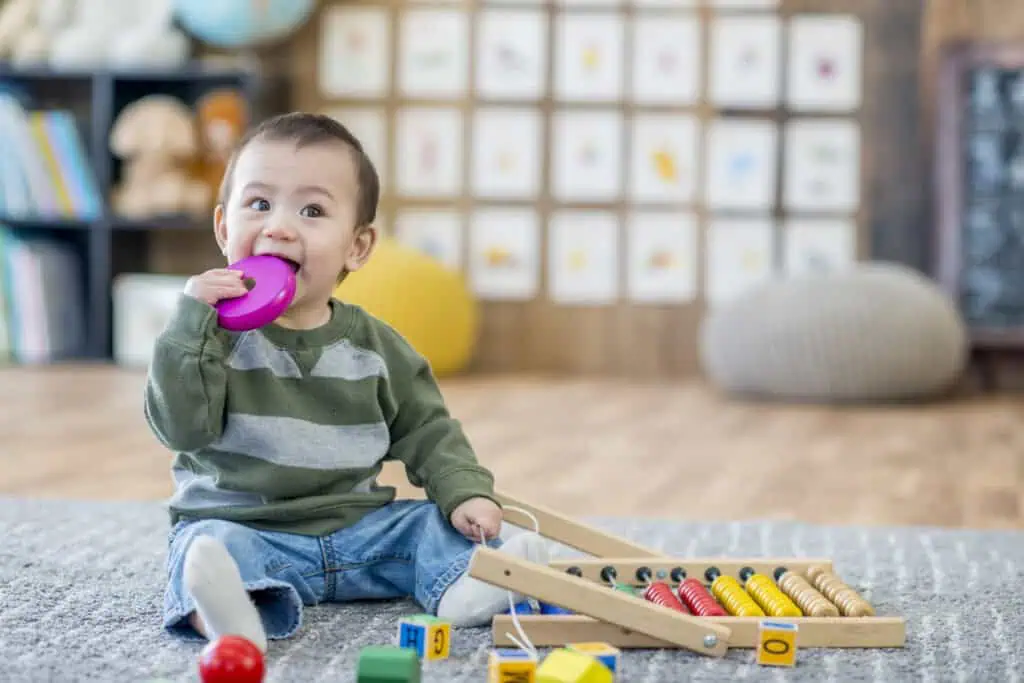 Um bebê com uma camisa listrada verde e cinza está sentado no chão acarpetado, mordiscando um brinquedo roxo. Vários brinquedos coloridos, incluindo um ábaco e blocos, estão espalhados diante deles. O ambiente aconchegante lembra os melhores ambientes de creche com fotos emolduradas e livros na estante ao fundo.