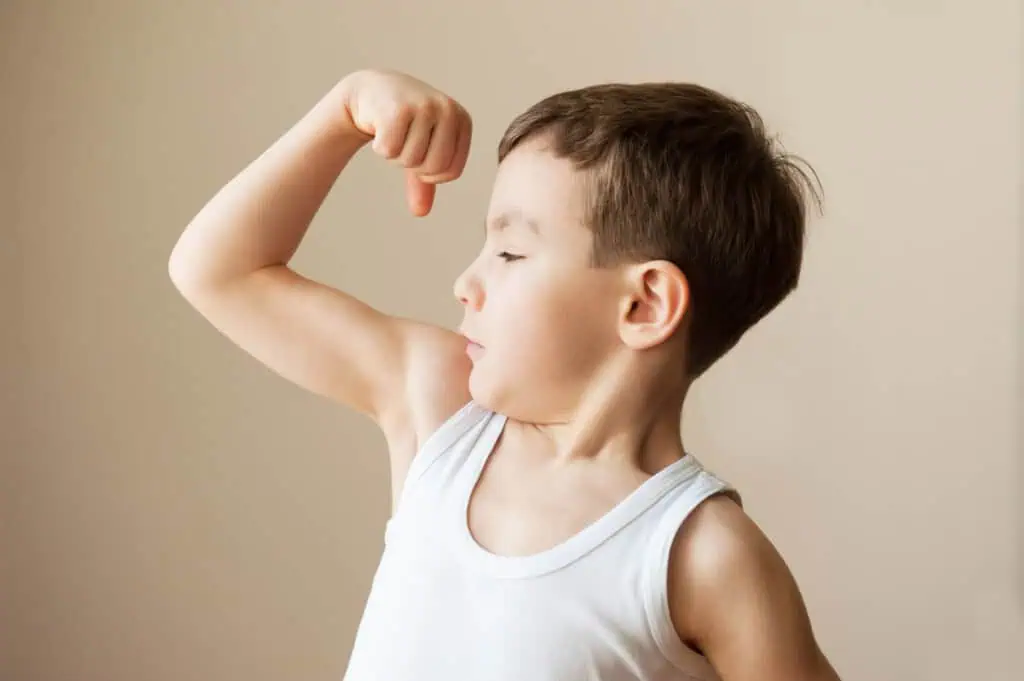 Um menino de camisa branca sem mangas flexiona o braço esquerdo, olhando para o bíceps com uma expressão determinada no rosto. O fundo é simples e neutro. Este momento poderoso é um lembrete de coisas sobre as quais nunca se deve provocar seu filho, pois a confiança dele é essencial para nutri-lo.