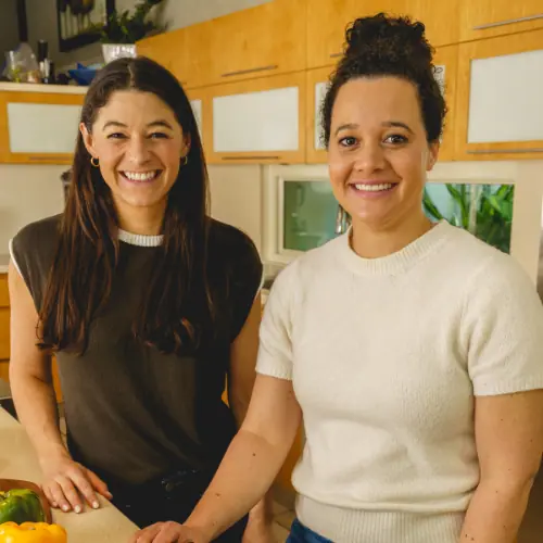 Duas pessoas (Ashley e Diana) estão em uma cozinha moderna. Uma tem longos cabelos escuros e veste uma camisa escura, enquanto a outra tem cabelos cacheados presos em um coque e veste um suéter leve. Eles estão sorrindo e parados perto de uma bancada com um pimentão.