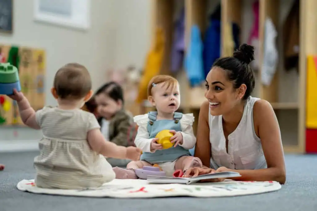 Uma mulher deitada num tapete, envolvendo-se alegremente com três crianças pequenas numa sala de jogos, personificando o calor que se espera encontrar ao considerar a melhor creche. Ela sorri para um bebê segurando um brinquedo amarelo, com prateleiras abertas de brinquedos e casacos proporcionando um cenário vibrante.