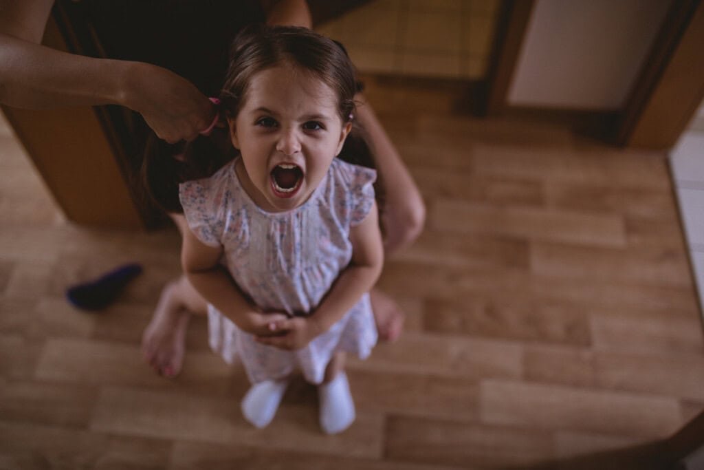 Uma jovem com um vestido floral está parada no chão de madeira com a boca aberta, parecendo estar gritando ou berrando. Um adulto, parcialmente visível, está atrás dela, penteando o cabelo da menina. Uma meia e uma escova de cabelo estão no chão próximas – como lidar com a conversa é evidente em sua dinâmica.