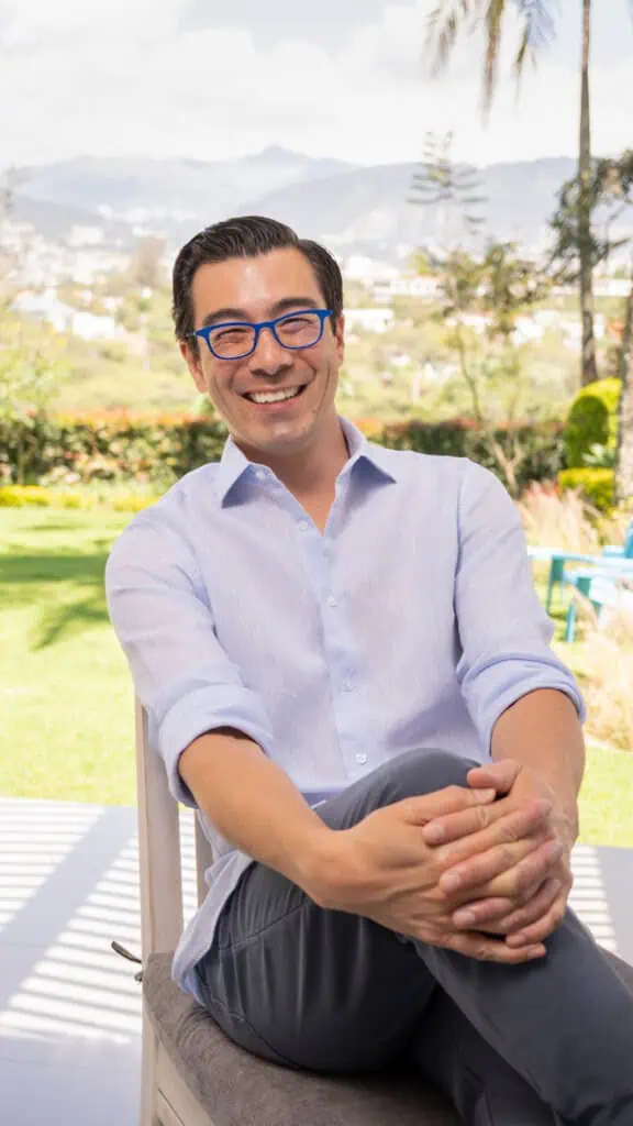 David Delisle vestindo uma camisa de botão azul claro e calças escuras está sentado em uma cadeira ao ar livre. Ele está sorrindo, com as pernas cruzadas e as mãos apoiadas no joelho. Ele também está usando óculos azuis. O fundo apresenta uma paisagem ensolarada com vegetação e colinas.