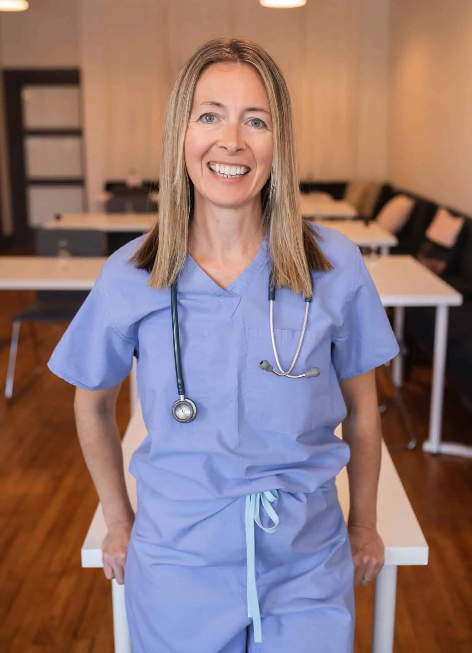 A Dra. Jessica Madden, de uniforme médico azul e com um estetoscópio no pescoço, sorri enquanto se apoia em uma mesa. A sala tem piso de madeira e mesas brancas com cadeiras pretas.