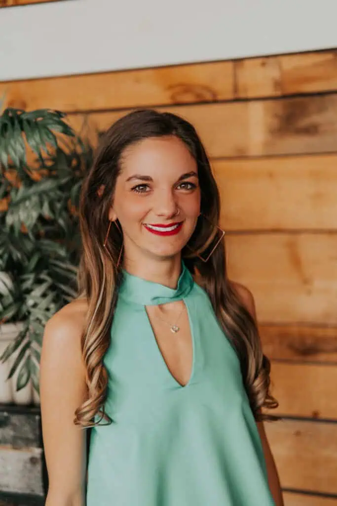 HeHe, tiro na cabeça de Stewart. Ela tem longos cabelos castanhos e usa uma blusa verde sem mangas. Ela está parada em frente a uma parede de madeira. Ela está sorrindo e usando grandes brincos de argola. Há uma planta ao fundo.
