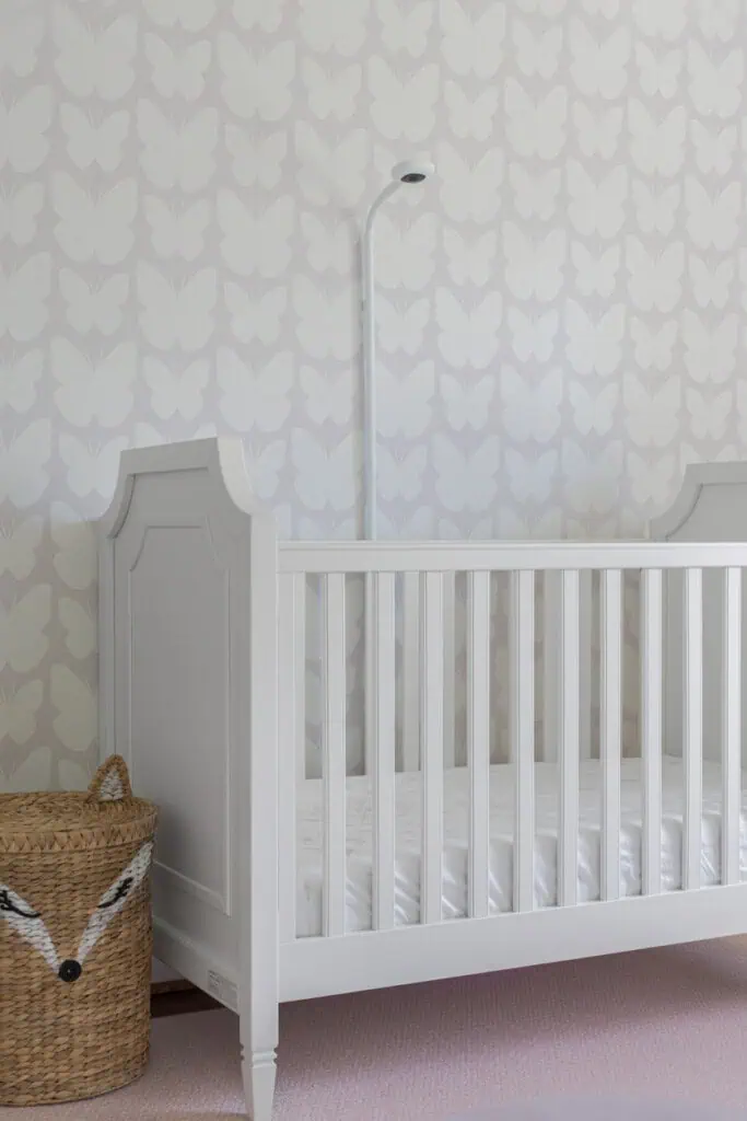 Um berço branco com design simples é colocado contra uma parede adornada com papel de parede com estampa de borboletas, refletindo as últimas tendências para bebês. À esquerda do berço há uma cesta de tecido exibindo uma cara de raposa caprichosa. Uma luminária de chão elegante fica próxima, criando um ambiente de berçário aconchegante e moderno.