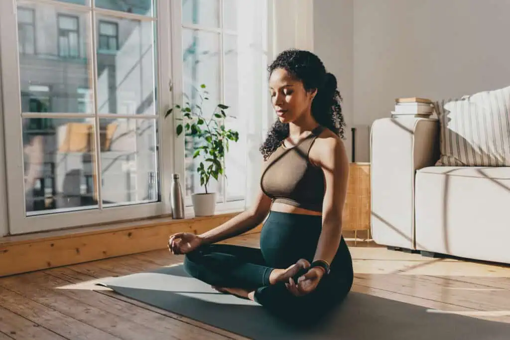 Uma mulher grávida está sentada em uma pose de ioga em uma esteira em uma sala iluminada pelo sol. Vestida com sutiã esportivo e leggings, seus olhos estão fechados em meditação. Uma grande janela, um vaso de plantas e um sofá com livros empilhados completam o ambiente sereno.