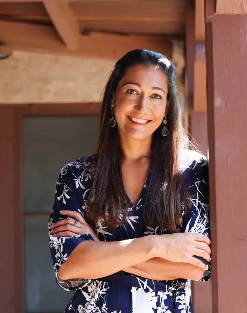 Alexandria DeVito, de cabelos longos, está com os braços cruzados, sorrindo, usando um vestido escuro com estampa floral. Ela está ao ar livre sob uma estrutura de madeira com vigas marrons e uma porta parcialmente visível ao fundo.