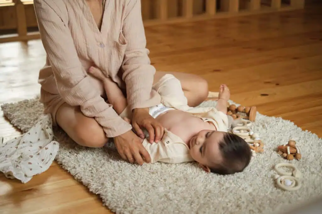 Uma mãe está sentada no chão, trocando a camisa de um bebê em meio a alguns brinquedos neutros. O bebê está deitado em um tapete branco com brinquedos de madeira espalhados por perto. O quarto, reflectindo um toque de estilo moderno de gravidez, apresenta pisos e paredes em madeira.