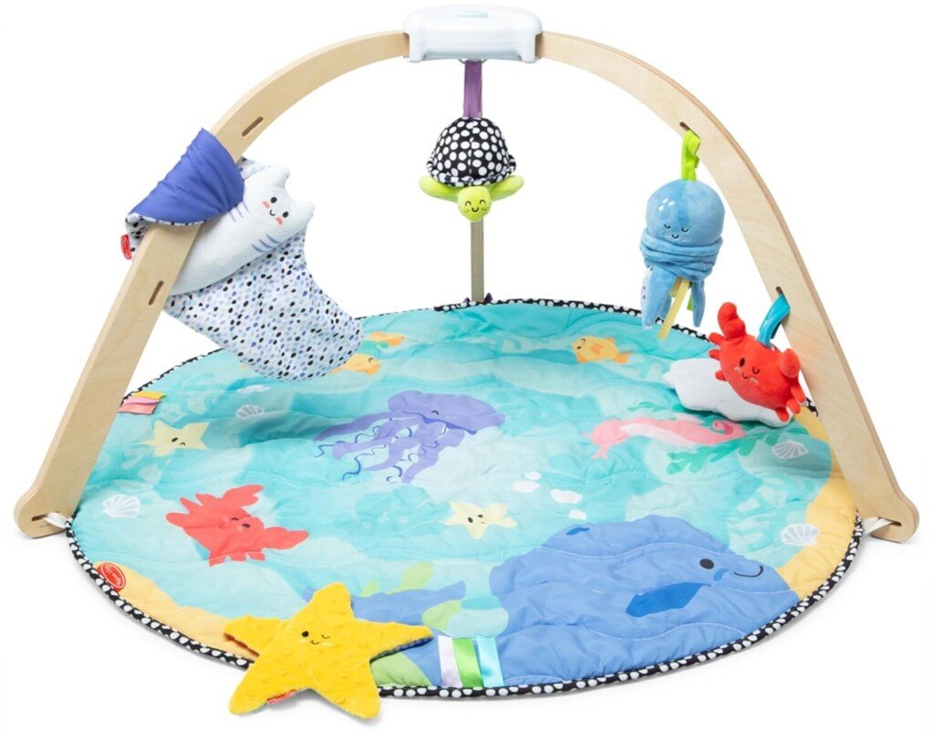 Um tapete colorido para brincar para bebês, parte de nossa coleção Produtos para bebês para 2025, apresenta uma moldura em arco de madeira com brinquedos pendurados, incluindo um urso branco de pelúcia e um polvo azul. O tapete redondo deslumbra com designs com temas marinhos, como estrelas do mar e conchas em um fundo azul sereno.