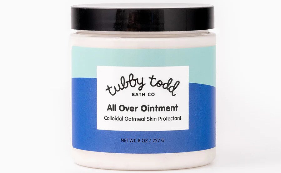 Um frasco de pomada Tubby Todd Bath Co All Over, rotulado como um produto essencial para bebês em 2025, apresenta um design azul e verde. Este recipiente de 8 onças (227 g) é um protetor de pele de aveia coloidal e vem com uma tampa preta elegante, colocada sobre um fundo branco liso.